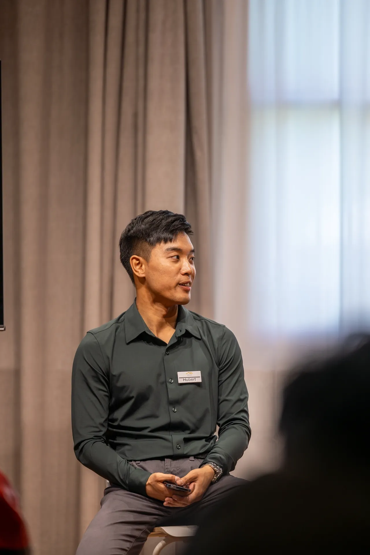 Man in dark shirt sitting indoors looking to the side.